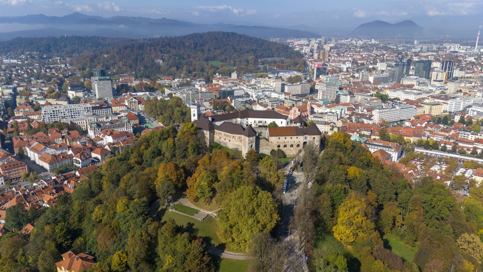 An aerial view of Ljubljana Castle on its hilltop above the terracotta rooftops of the city, with the Julian Alps in the distance